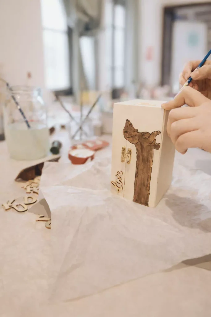 Dłonie malujące pędzlem brązową postać psa na jasnym, drewnianym pudełku. Obok leżą wycięte z drewna litery i słoik z wodą do pędzli.