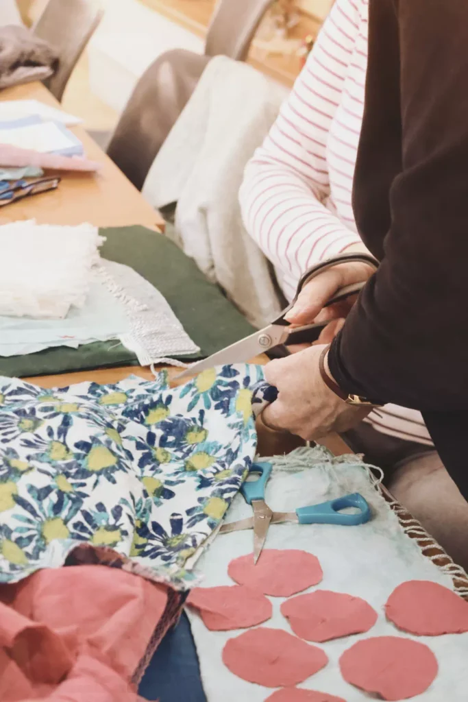 Zbliżenie na dłonie wycinające wzorzysty materiał w kwiaty dużymi nożyczkami krawieckimi. Na stole leżą różowe skrawki tkaniny i niebieskie nożyczki.
