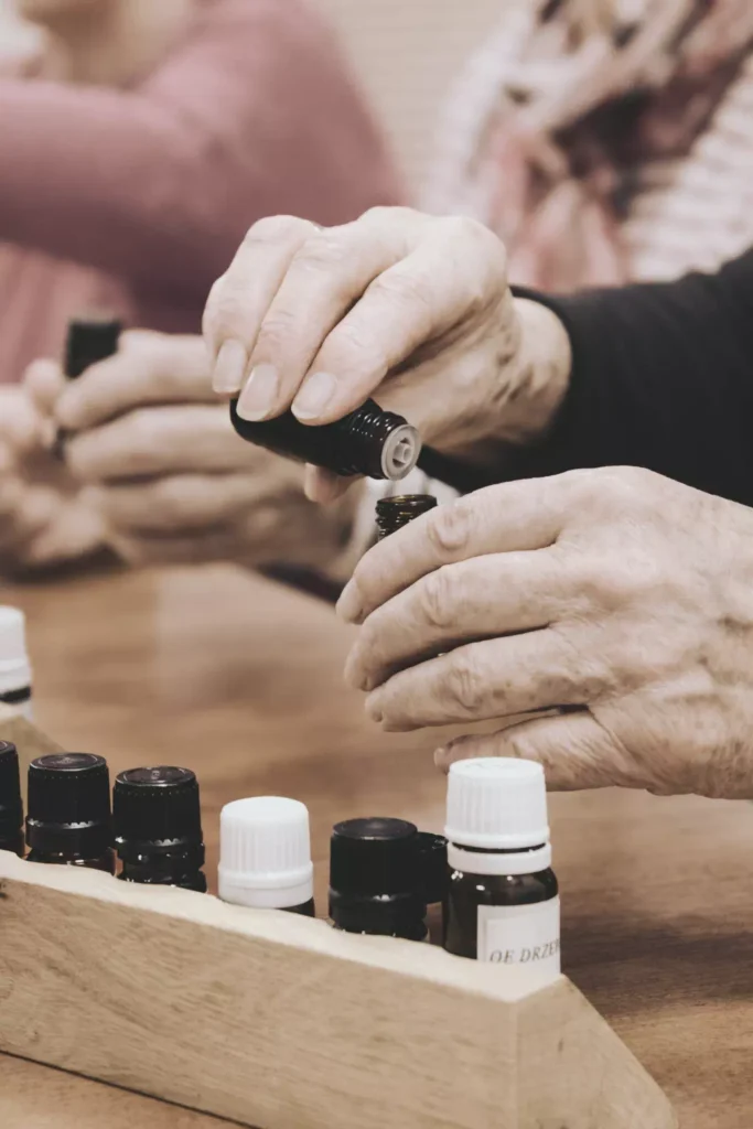 Zbliżenie na dłonie seniorki przelewającej olejek eteryczny z małej buteleczki do większego flakonu podczas warsztatów.