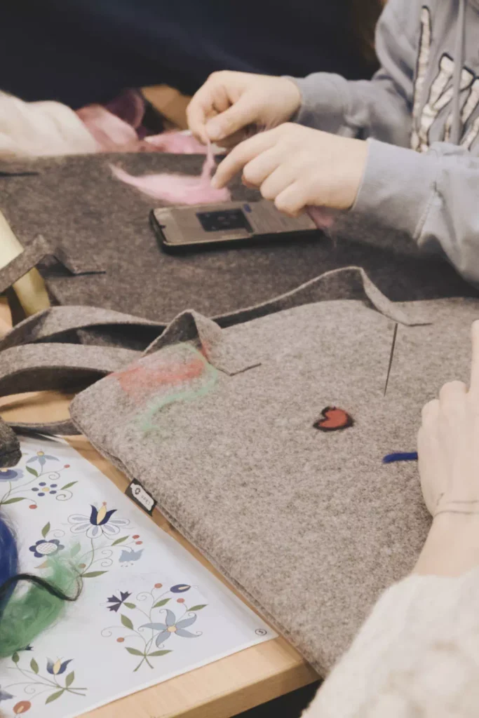 Dłonie uczestników warsztatów filcujących kolorowe wzory (serce i motywy roślinne) na szarej torbie filcowej przy użyciu igły i wełny czesankowej.