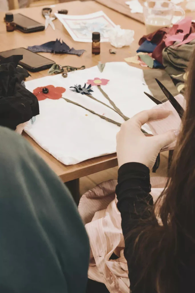 Warsztaty z tworzenia kwiatowych kompozycji z materiału. Na pierwszym planie dłonie z nożyczkami, na stole gotowy wzór trzech kwiatów na białym tle.