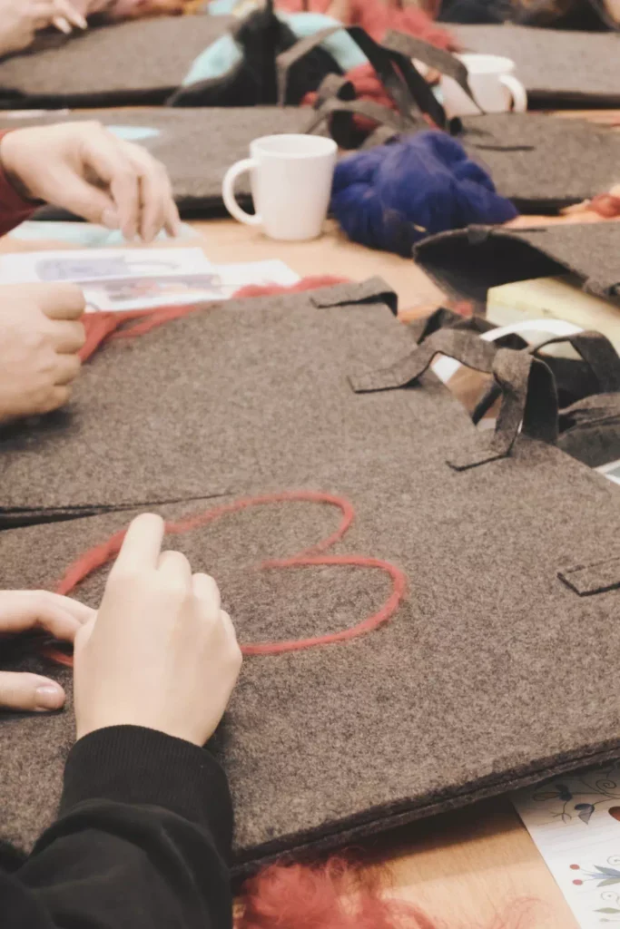 Personalizowanie filcowej torby – dłonie dobierające różową wełnę czesankową do stworzenia aplikacji na szarym materiale.