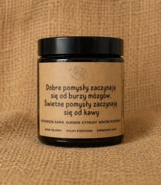 Świeca sojowa w szklanym, brązowym słoiku z etykietą z napisem "Dobre pomysły zaczynają się od burzy mózgów. Świetne pomysły zaczynają się od kawy" o słodkim, kawowym zapachu.