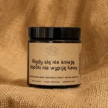 Świeca sojowa w szklanym, brązowym słoiku z etykietą z napisem "Nigdy się nie śmieję, dopóki nie wypiję kawy." o słodkim, kawowym zapachu.