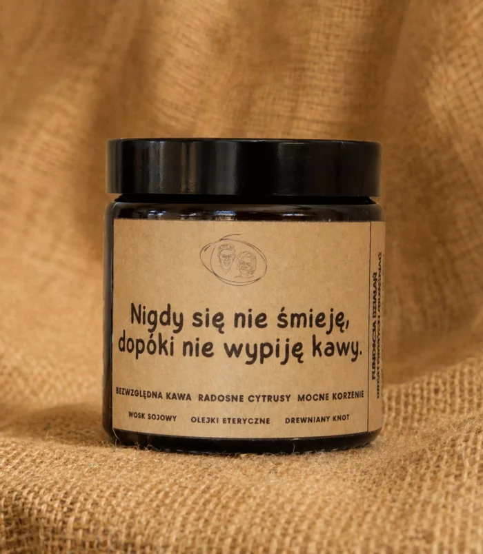 Świeca sojowa w szklanym, brązowym słoiku z etykietą z napisem "Nigdy się nie śmieję, dopóki nie wypiję kawy." o słodkim, kawowym zapachu.