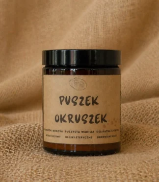 Świeca sojowa w szklanym słoiku w kolorze brązowym z etykietą z napisem "Puszek okruszek" o słodkim, relaksującym zapachu.
